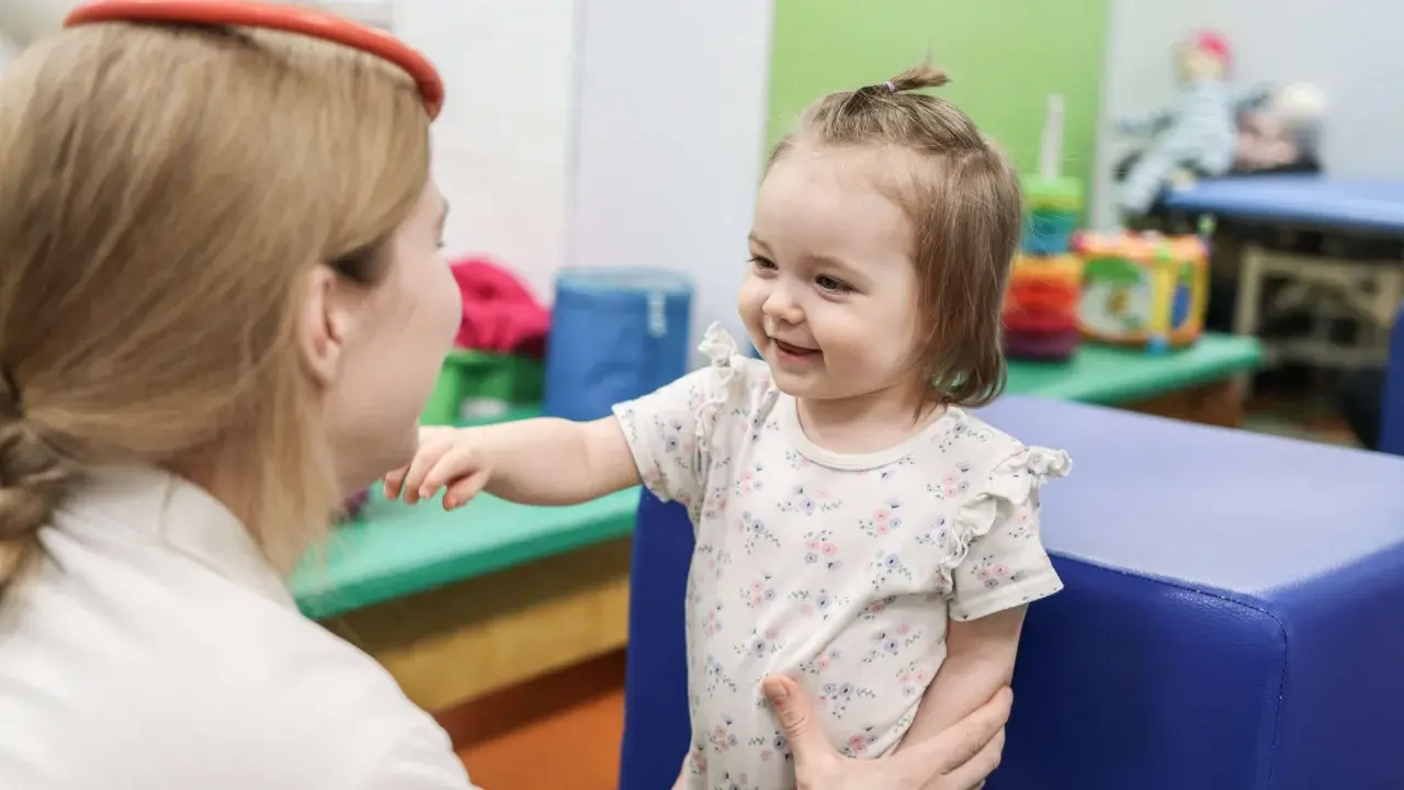 Uśmiechnięte dziecko w białej bluzce z kwiatkami wyciąga rączkę do terapeuty.
