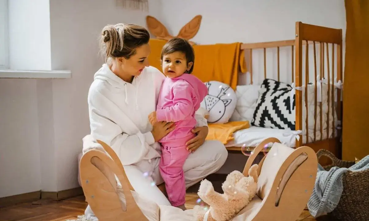 Kobieta w białej bluzie trzyma dziecko w różowym dresie. W tle łóżeczko i drewniana huśtawka z misiem.