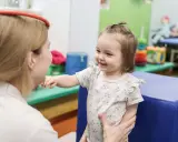 Uśmiechnięte dziecko w białej bluzce z kwiatkami wyciąga rączkę do terapeuty.
