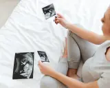 Kobieta w ciąży ogląda zdjęcia USG płodu na białym łóżku.