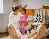 Kobieta w białej bluzie trzyma dziecko w różowym dresie. W tle łóżeczko i drewniana huśtawka z misiem.