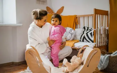 Kobieta w białej bluzie trzyma dziecko w różowym dresie. W tle łóżeczko i drewniana huśtawka z misiem.