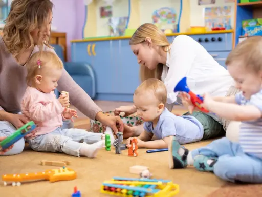 Opiekunki bawią się z dziećmi zabawkami na dywanie w żłobku.