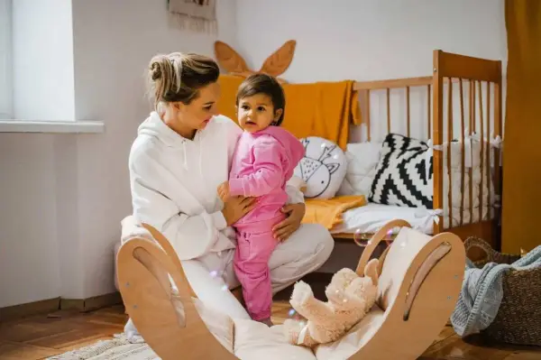 Kobieta w białej bluzie trzyma dziecko w różowym dresie. W tle łóżeczko i drewniana huśtawka z misiem.